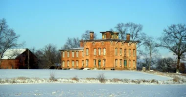 A front view of Prospect Place showing tall white columns, a large red-brick facade, symmetrical windows, and a wide front porch under a clear sky.