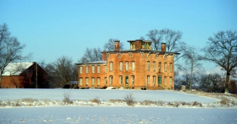 A front view of Prospect Place showing tall white columns, a large red-brick facade, symmetrical windows, and a wide front porch under a clear sky.