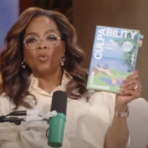 Oprah Winfrey holding up the book The Culpability by Holsinger while speaking into a microphone.